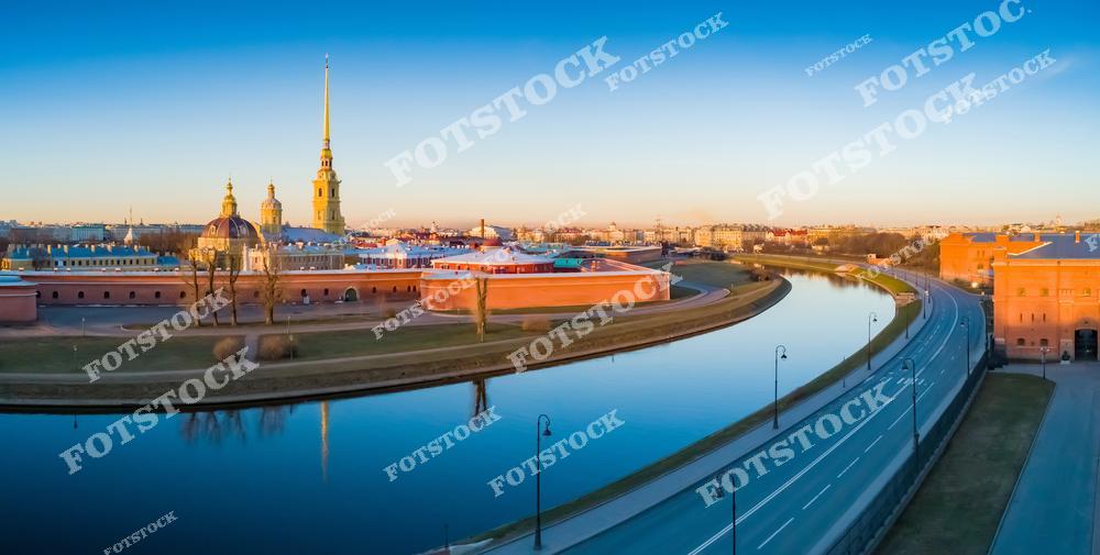На фотографии изображена Петропавловская крепость, одна из главных достопримечательностей Санкт-Петербурга. Вид на Кронверкский пролив с ровным зеркальным отражением крепостных стен. На заднем плане видны исторические здания и городская панорама. Закатное освещение придаёт теплые оттенки городскому пейзажу.
